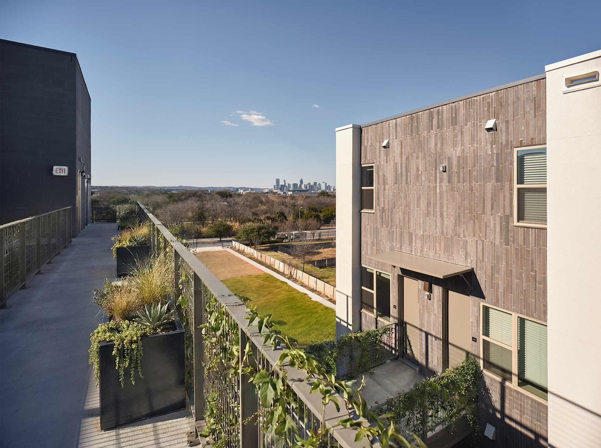 Elevated walkway with planters between modern apartments featuring wood-panel cladding and city