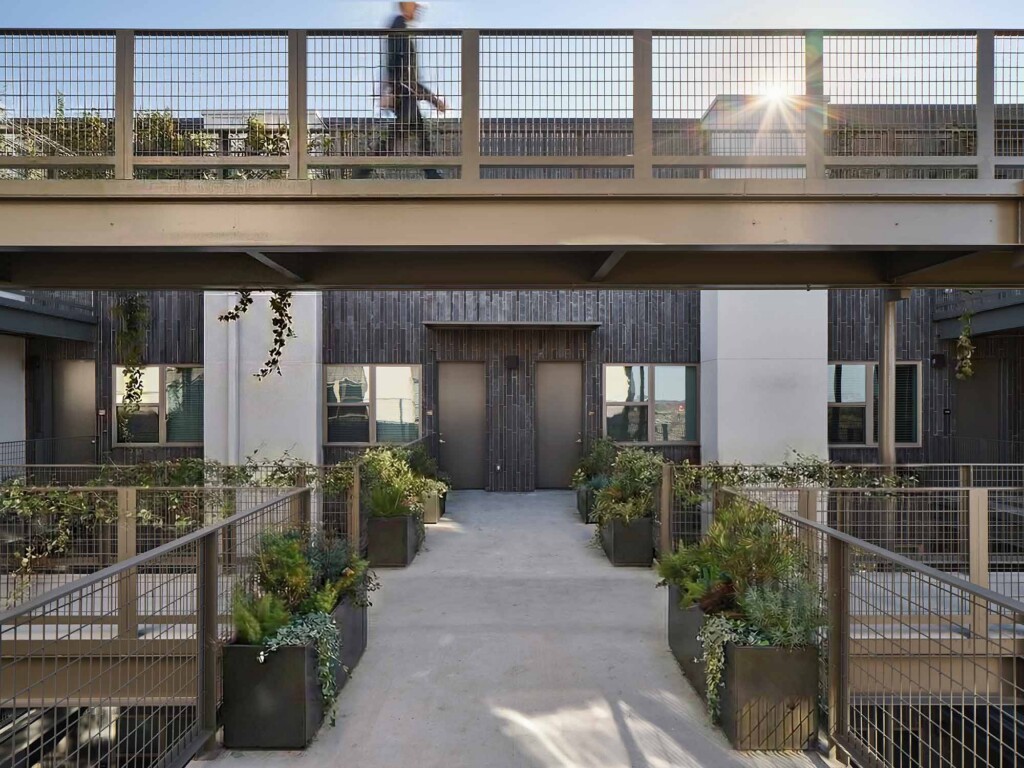 Courtyard walkway with planters leading to dark brick building entrance, person crossing bridge