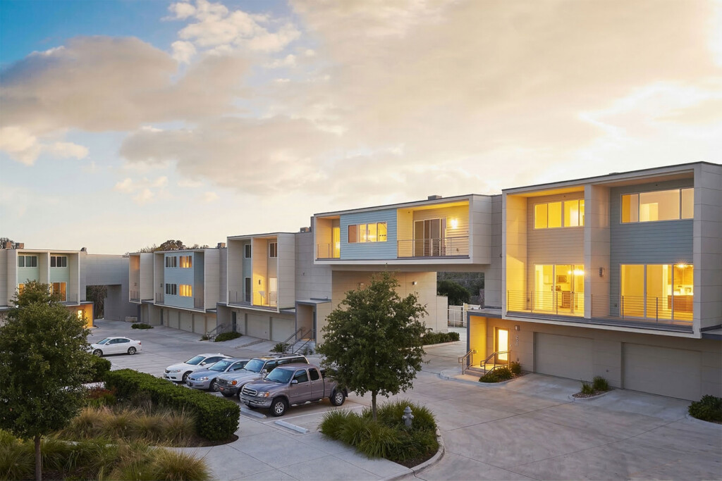 Modern townhouse complex at dusk with gray siding, large windows, balconies, parking lot and skybridge