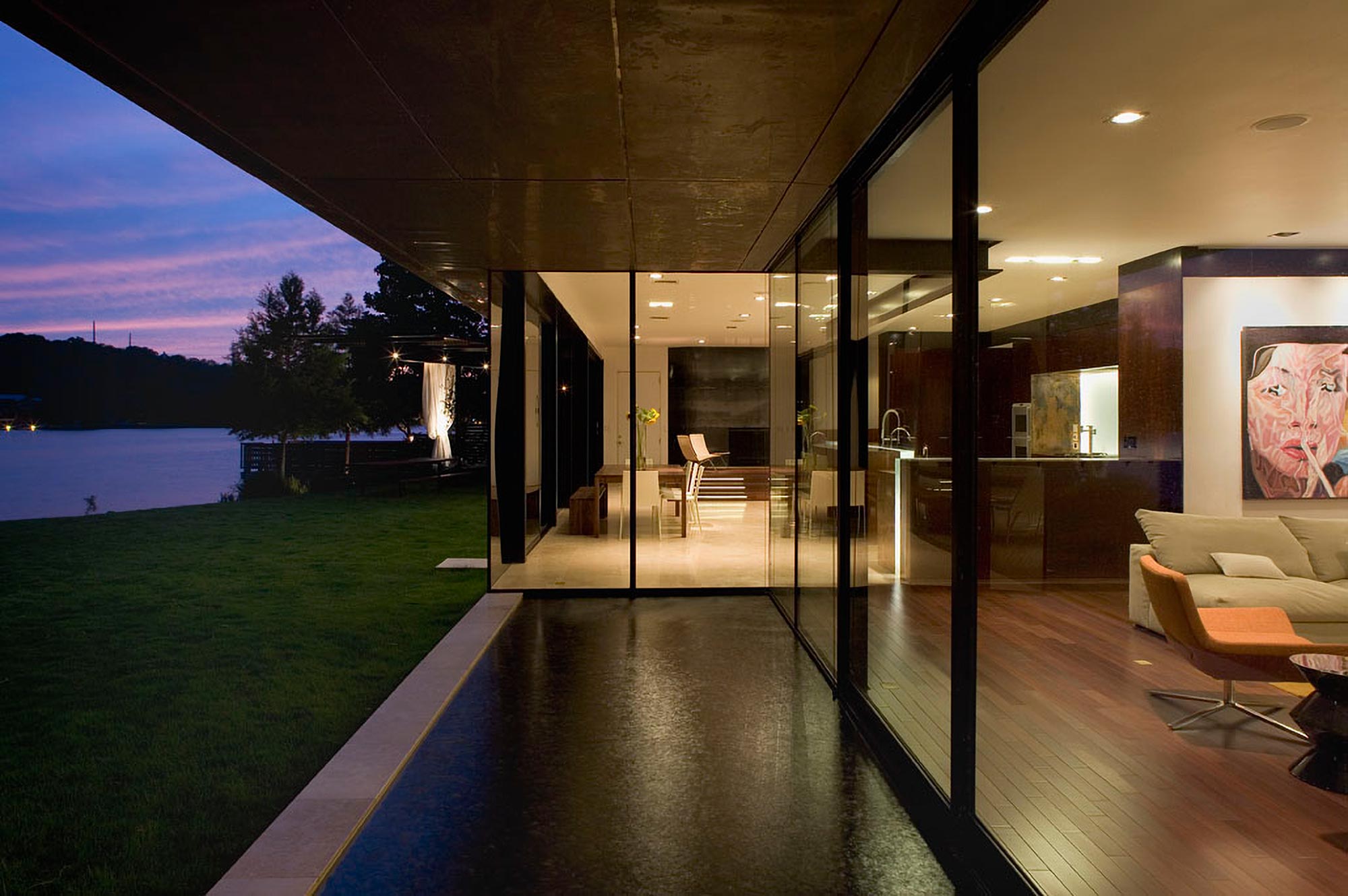 Modern glass-walled lakeside home at dusk with open living space, concrete ceiling, and lawn