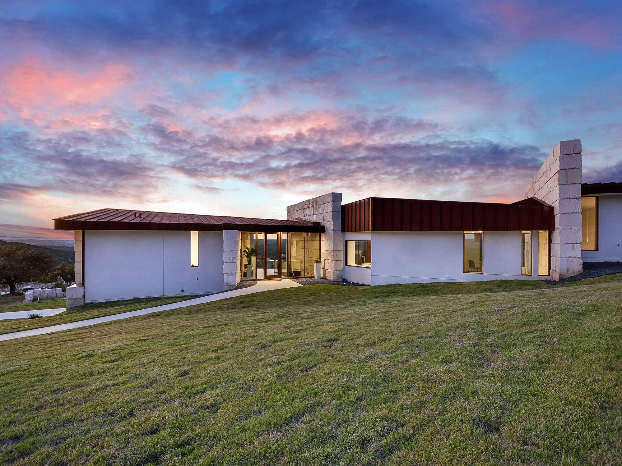 Modern single-story home with white stucco walls stone columns and copper roof at sunset with green
