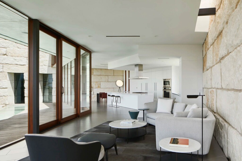 Open plan living area with stone walls wood-framed glass doors white kitchen and gray sectional sofa