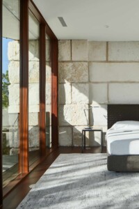 Modern bedroom with limestone block wall, wood-framed glass doors, gray rug, and minimalist bed