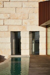 Stone wall facade with two narrow windows, wooden deck and pool, corten steel overhang above