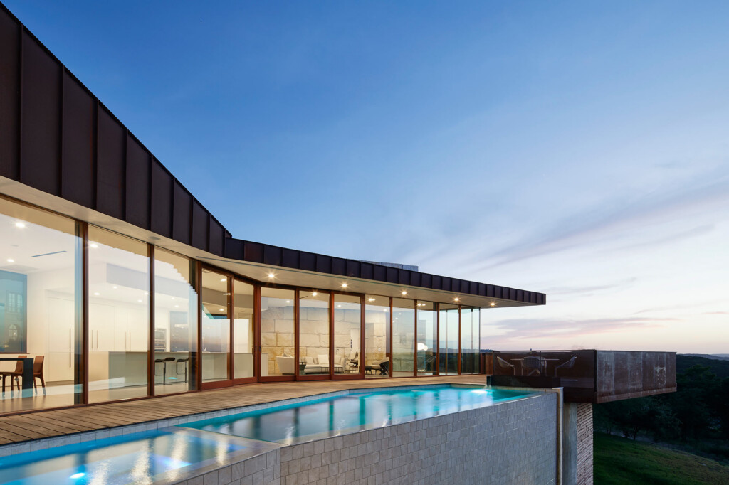 Modern hillside home with dark metal roof, floor-to-ceiling glass walls, wood deck and infinity pool