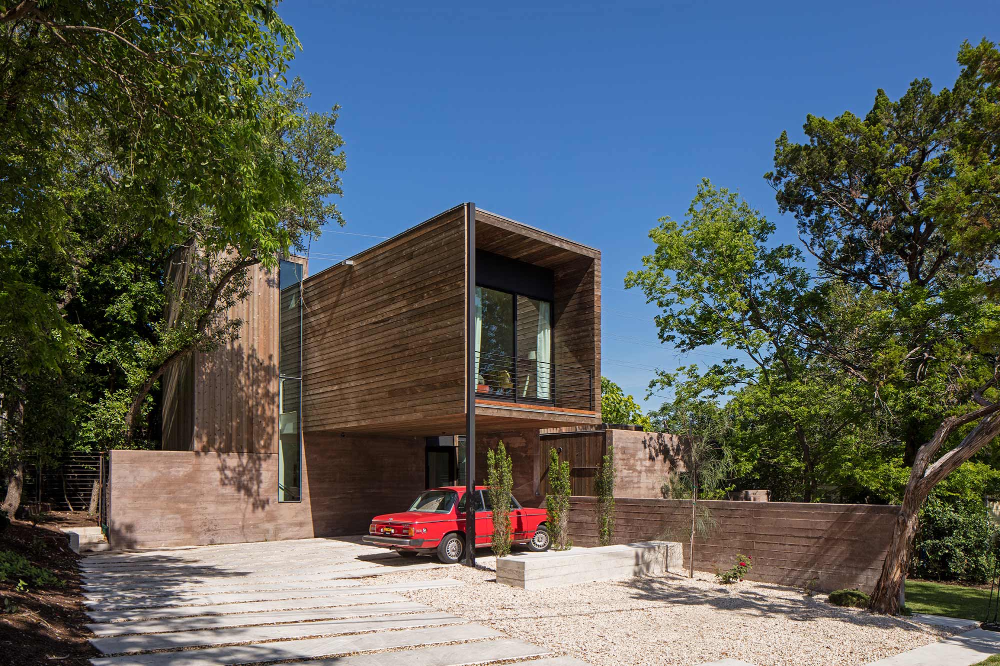 Modern two-story wood-clad house with cantilevered upper volume, rammed earth walls, and red