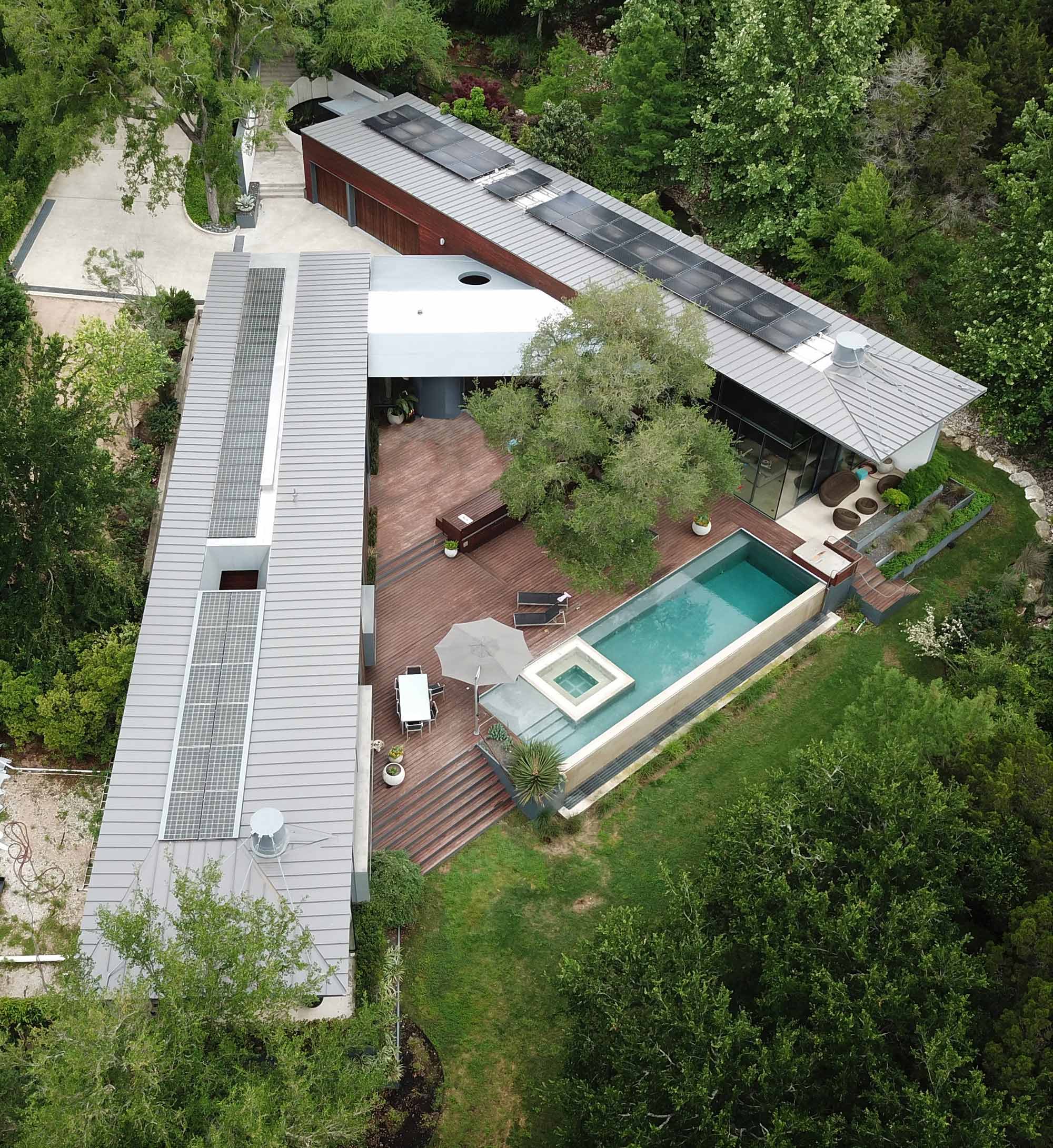 Aerial view of modern L-shaped home with metal roof solar panels wood deck pool and lush greenery