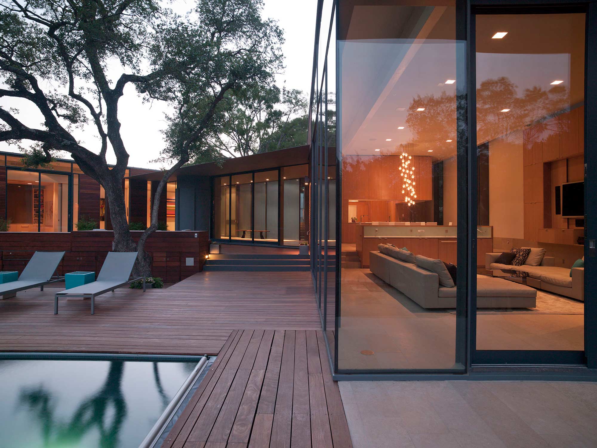 Modern home with wood deck courtyard, pool edge, mature oak tree, and glass walls revealing living