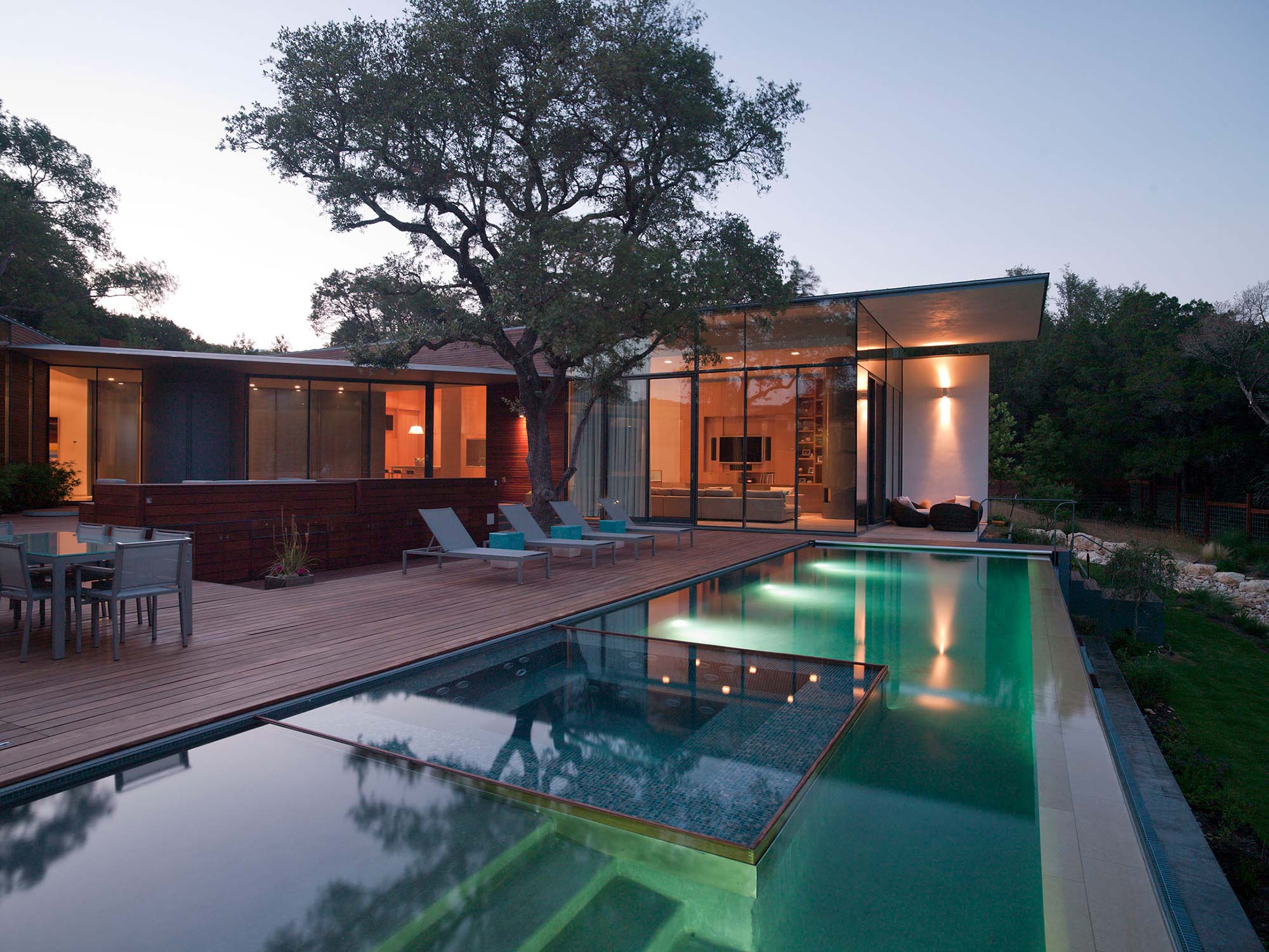 Modern glass pavilion home at dusk with illuminated pool, wood deck, lounge chairs and mature oak