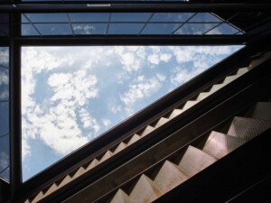 Upward view of metal staircase with perforated treads against glass skylight and blue cloudy sky