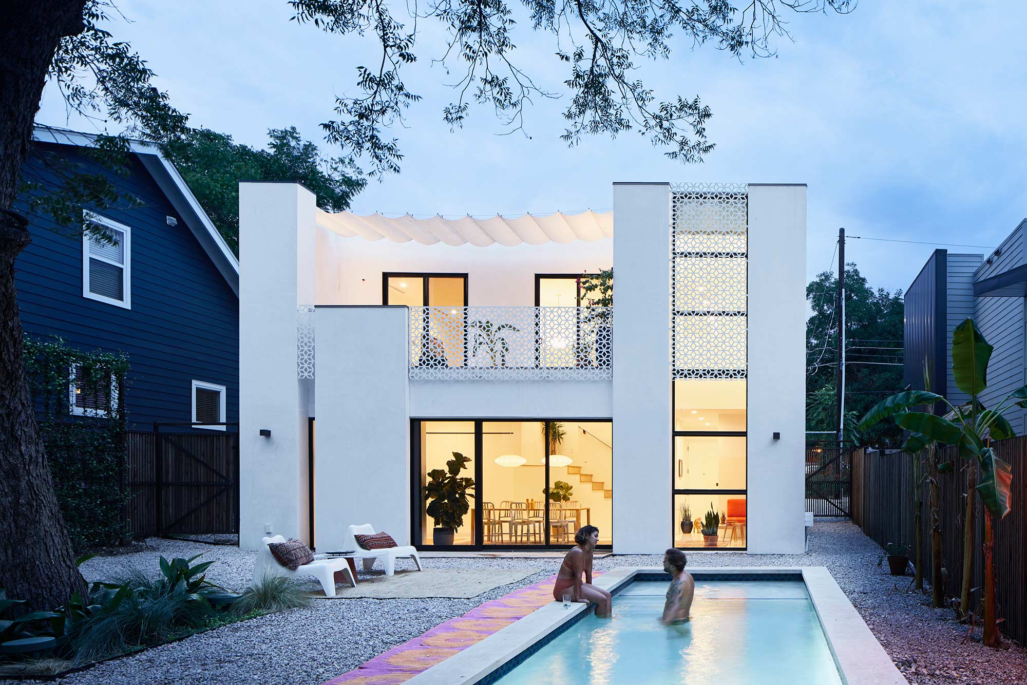 Modern white stucco home with decorative screens, large windows, pool with two people, gravel yard