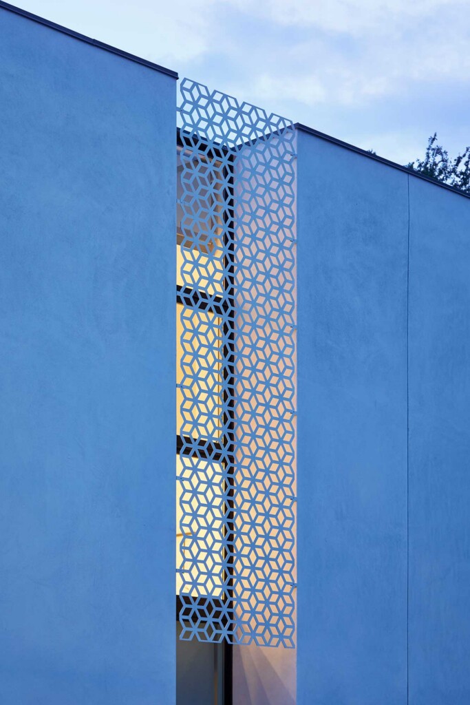 Blue stucco building facade with tall vertical window featuring geometric metal screen pattern