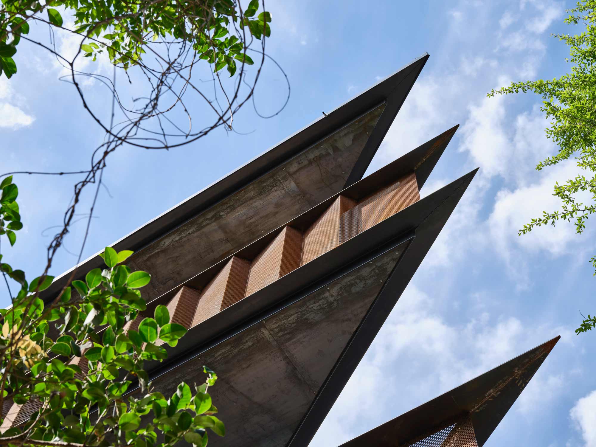 Angular dark metal facade with triangular projections and copper mesh screens framed by green