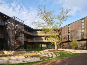 Modern courtyard with corten steel and dark metal buildings, external staircase, mature tree, and