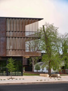 Modern building with weathered corten steel cladding and glass walls beside mature trees and white