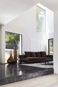 Modern living room with dark sofa on polished concrete platform, tall windows, white walls, bronze