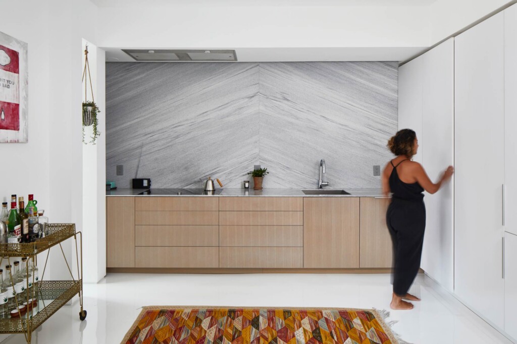 Woman opening tall cabinet in minimalist kitchen with marble backsplash and oak cabinetry
