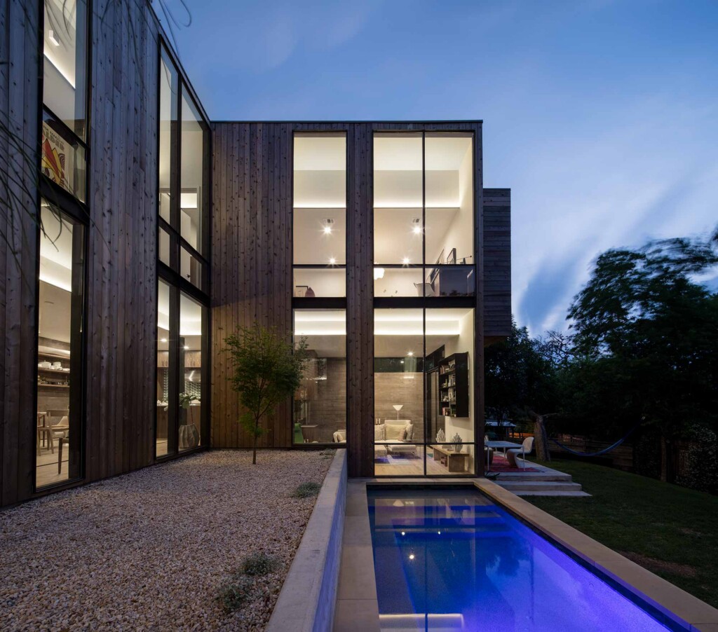 Modern two-story home with dark wood cladding, floor-to-ceiling windows, gravel courtyard and lap
