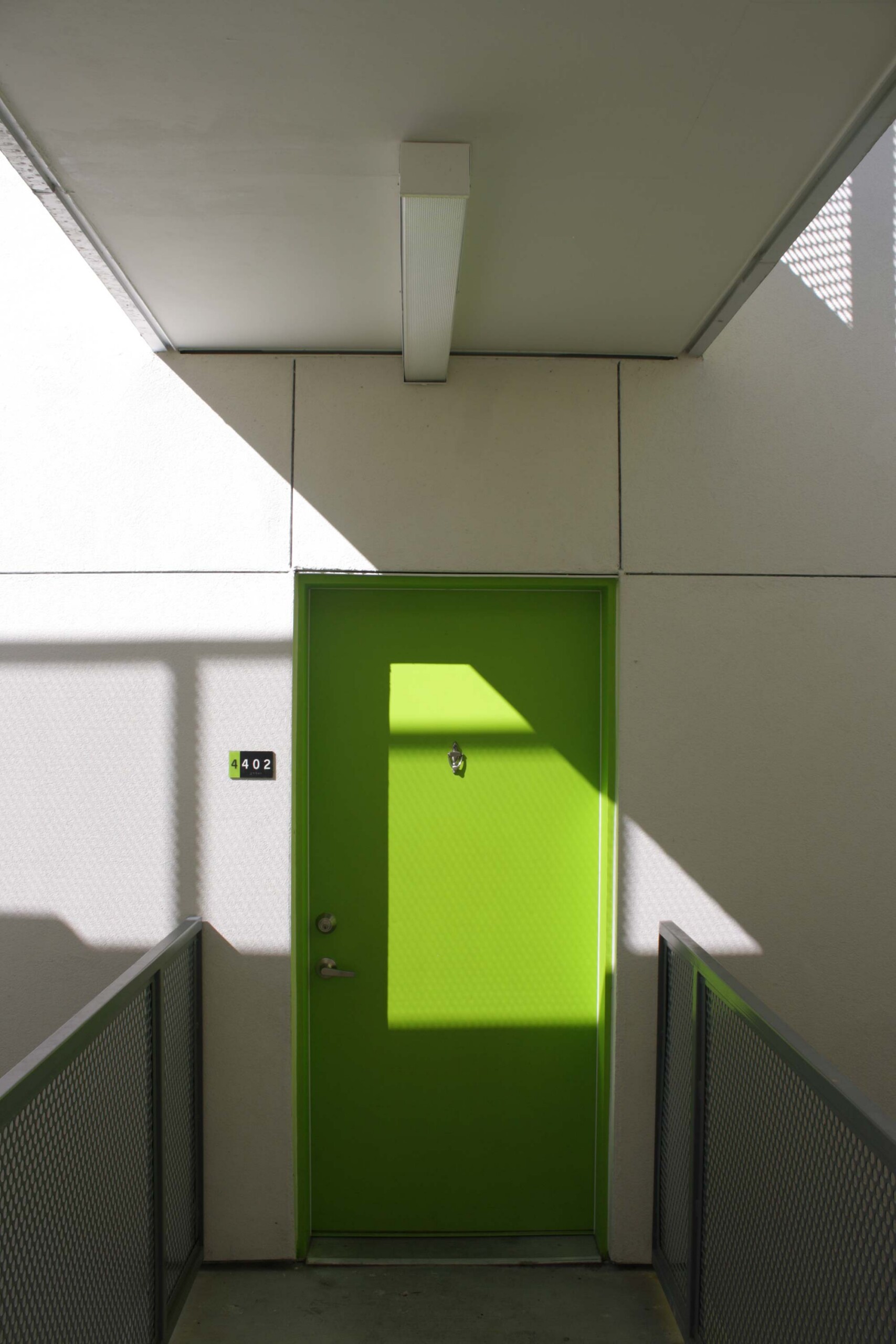 Bright lime green door numbered 4402 in white concrete corridor with metal railings and sunlight
