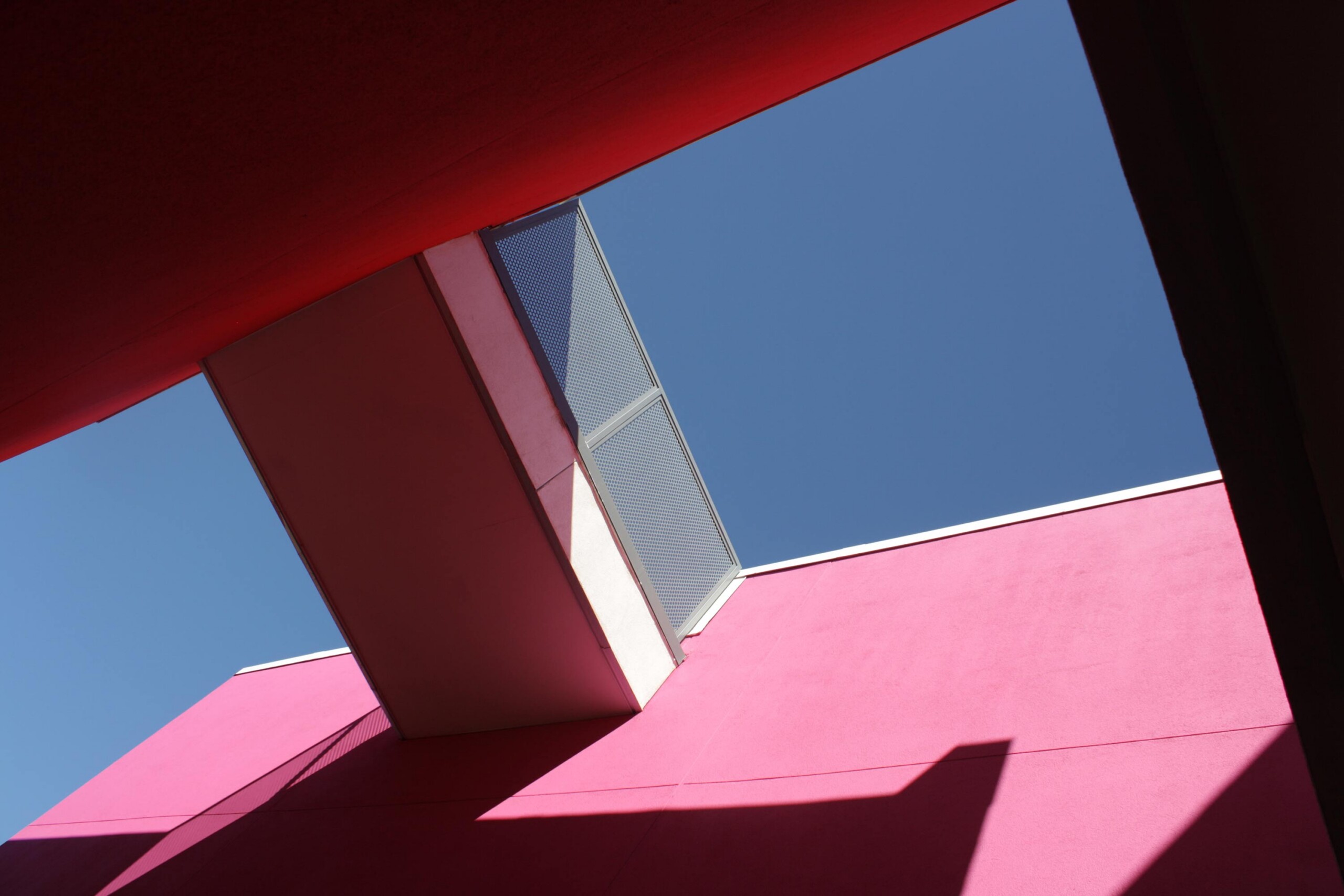 Abstract view of pink and red geometric walls against blue sky with angular shadows and mesh screen