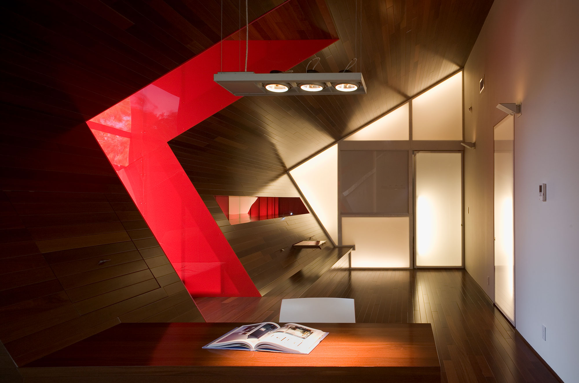 Angular red sculptural element in wood-paneled interior with dramatic lighting and open book on desk