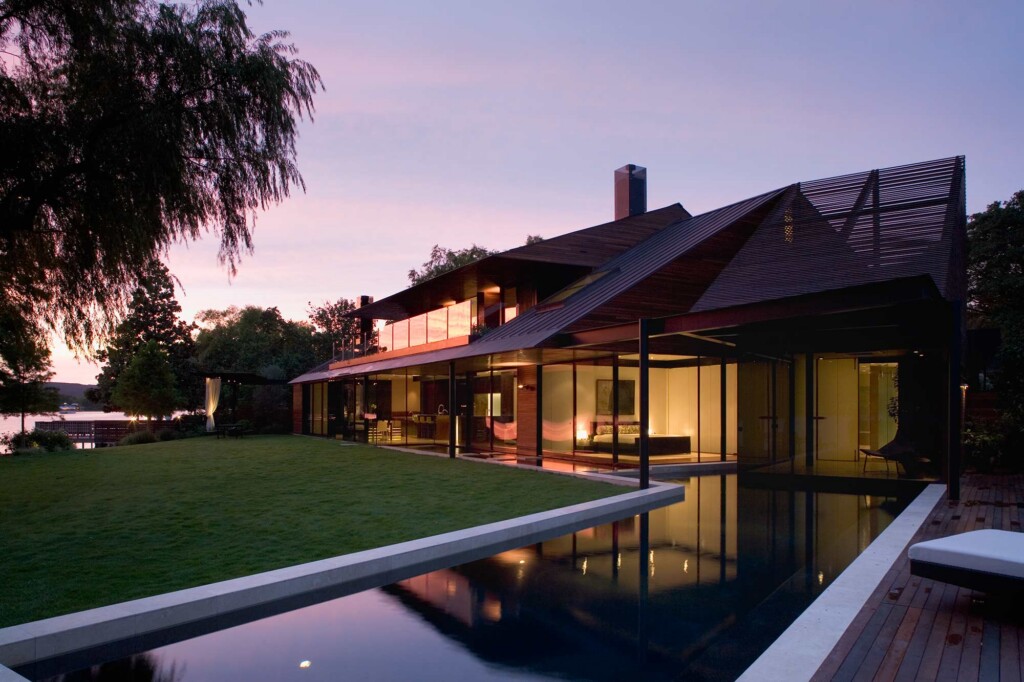 Modern lakeside home at dusk with glass walls, metal roof, manicured lawn, and reflective lap pool
