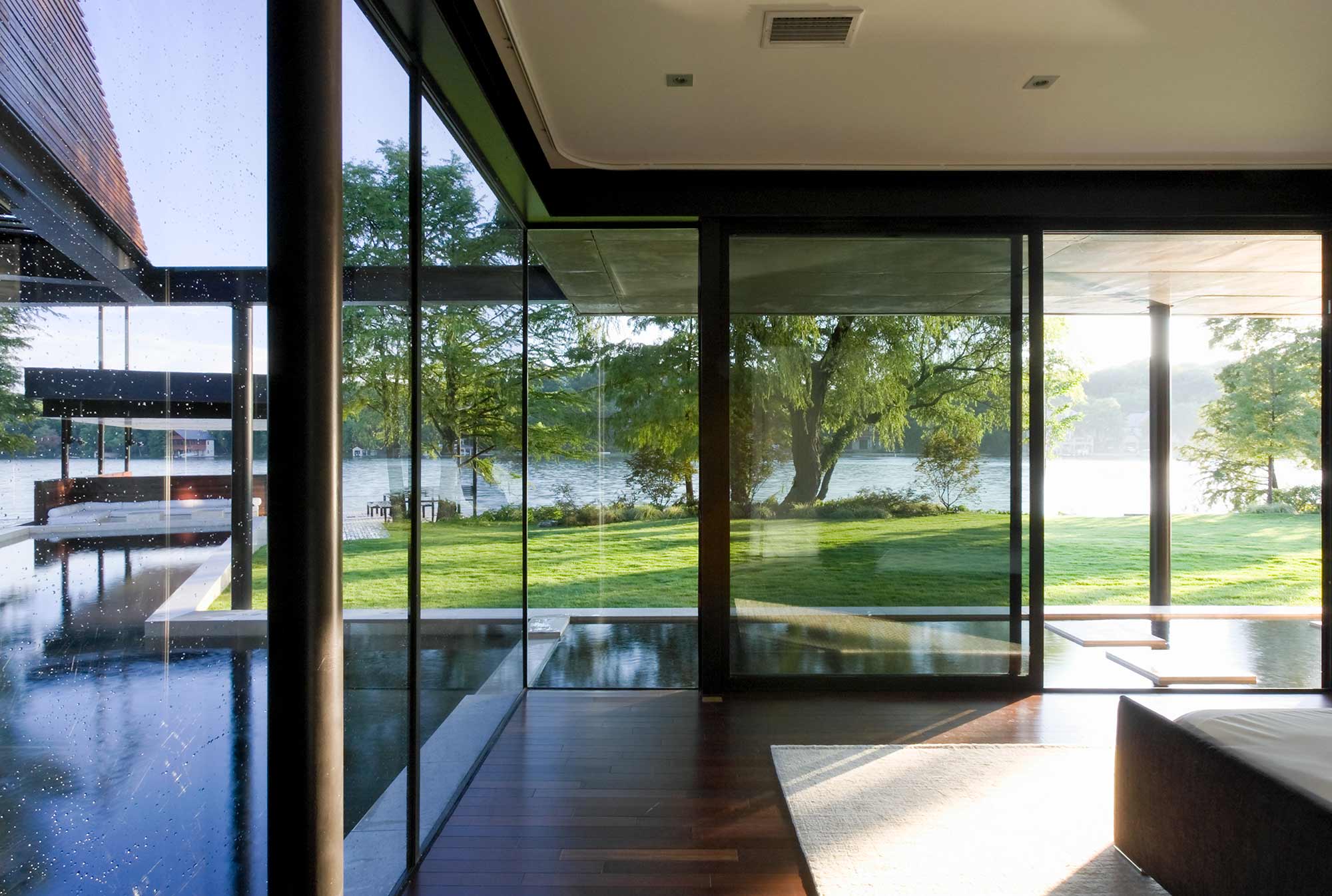 Modern glass-walled interior with dark wood floors overlooking manicured lawn and lake with trees