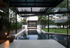 Modern glass-enclosed pool pavilion with steel frame overlooking lake and manicured lawn at dusk
