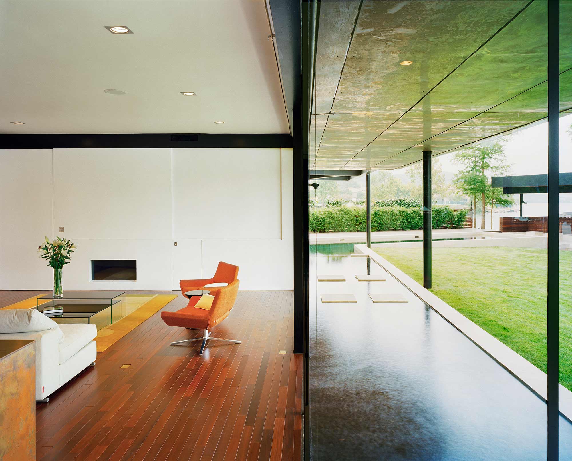 Modern living room with orange chair, wood floors, white walls opening to pool and lawn via glass
