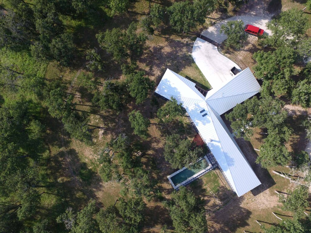 Aerial view of cross-shaped ranch house with white metal roof surrounded by trees and a pool