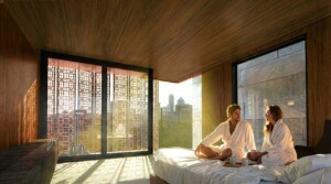 Couple in white robes on bed in wood-paneled hotel room with geometric screen and city view