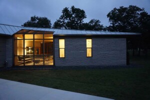 Modern ranch house at dusk with stone walls, metal roof, and illuminated glass-enclosed sunroom