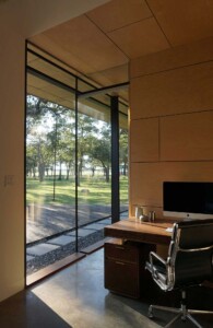 Modern home office with floor-to-ceiling glass walls, wood-paneled ceiling, desk with iMac, Eames