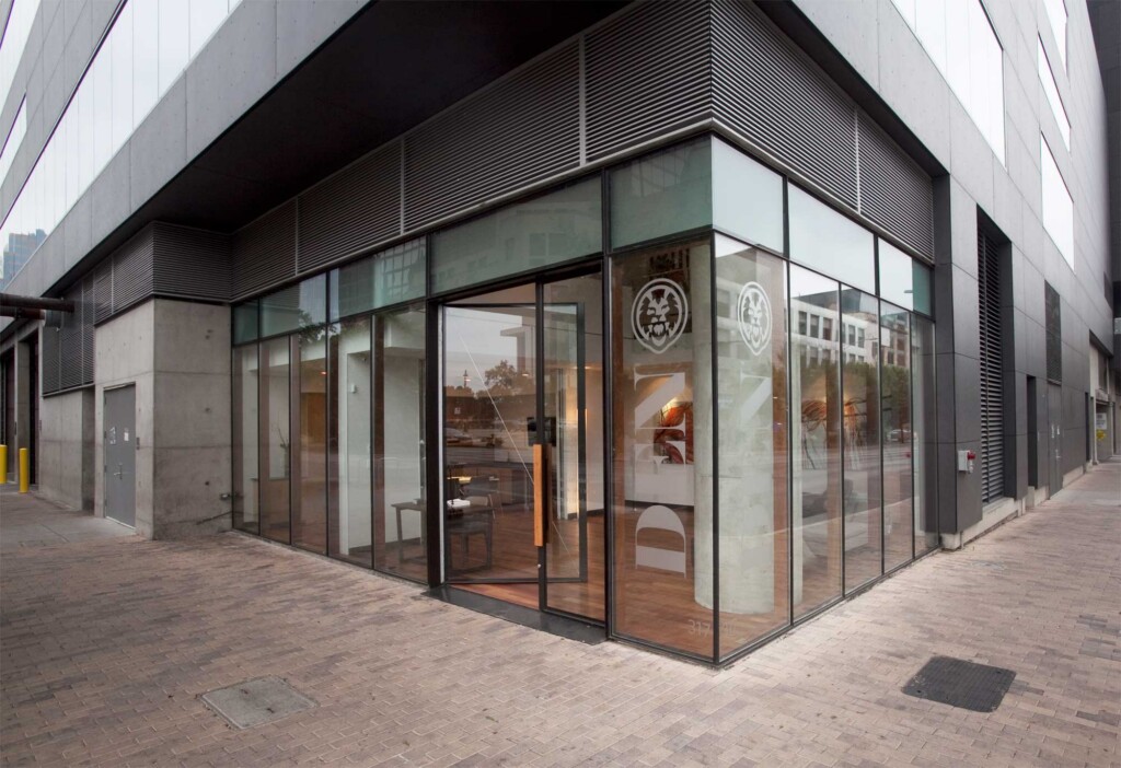 Corner view of modern glass-fronted retail space with DEN logo, black metal framing, and brick