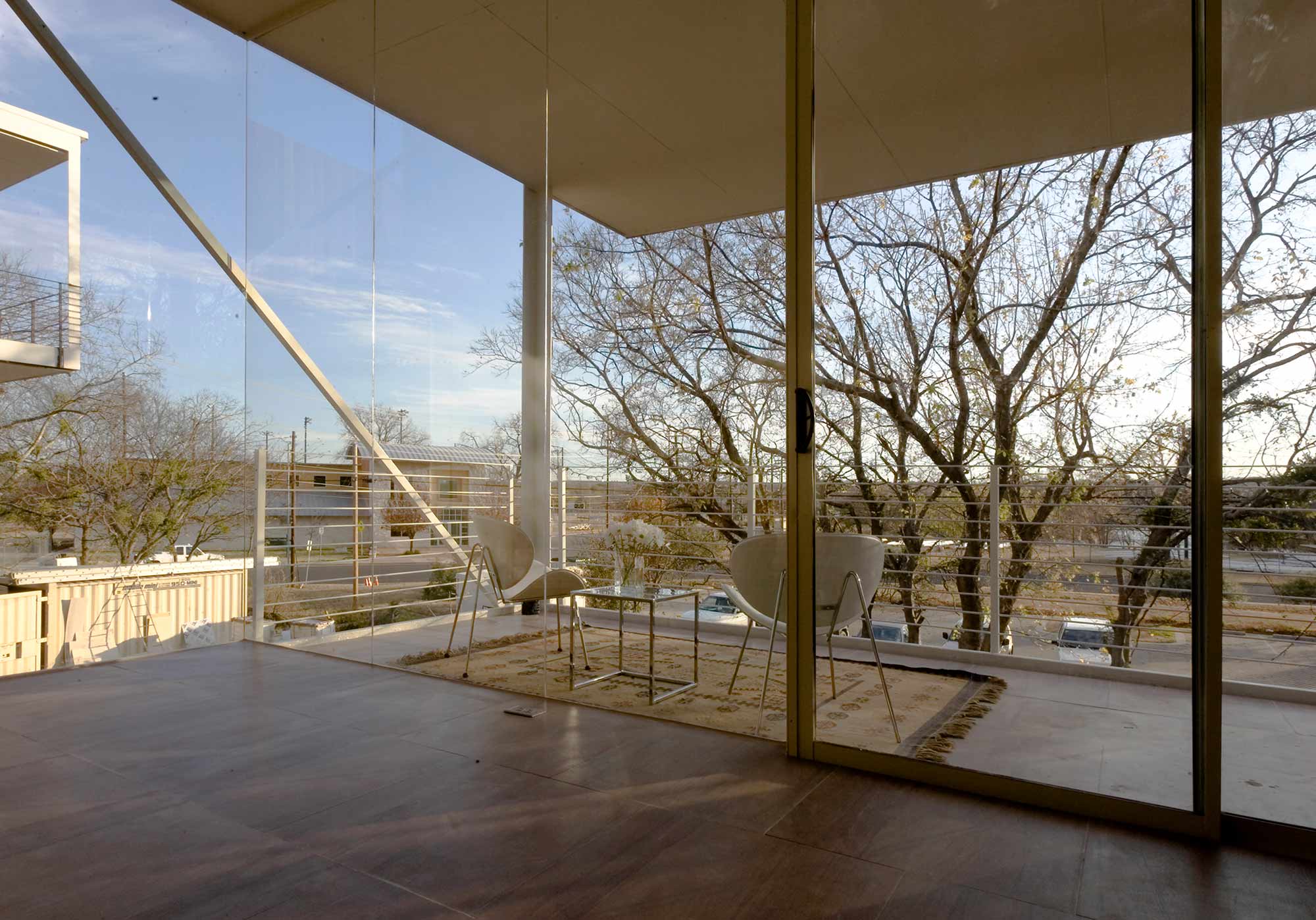 Glass-walled modern interior with floor-to-ceiling windows opening to balcony with two white chairs