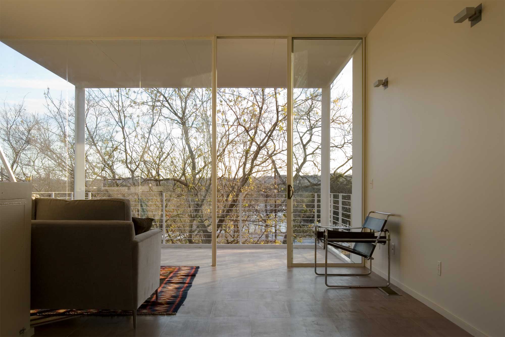 Minimalist living room with floor-to-ceiling windows, gray sofa, Wassily chair, and tree views